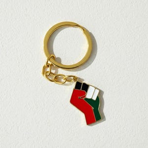 Peut inclure: Un porte-clés doré avec un charm en forme de poing. Le poing est coloré avec de l'émail rouge, noir, blanc et vert. Le porte-clés a un anneau rond et une petite chaîne reliant le charm.
