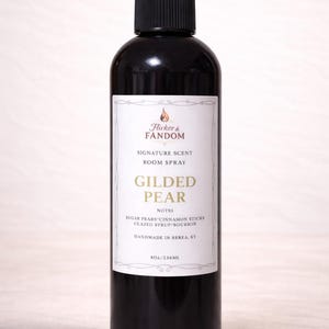 May include: A black room spray bottle with a white label featuring the text "GILDED PEAR" and the brand name "Flicker & Fandom." The label lists notes of sugar pears, cinnamon sticks, and glazed syrup/bourbon. The bottle contains 8 oz / 236 ml.