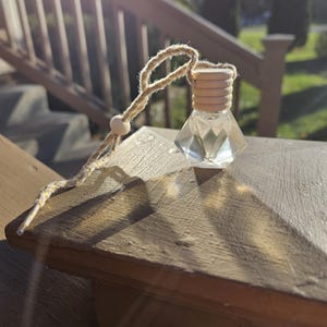 May include: A clear, faceted glass bottle with a wooden cap and a woven rope loop. The bottle is hanging on a wooden surface. The background includes a wooden railing and trees.