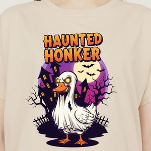 Könnte beinhalten: Beiges T-Shirt mit einem Halloween-Motiv. Das Design zeigt eine Cartoon-Ente in einem Geisterkostüm, mit dem Text "HAUNTED HONKER". Der Hintergrund zeigt eine gruselige Szene mit lila Himmel, Fledermäusen und Spukhäusern.