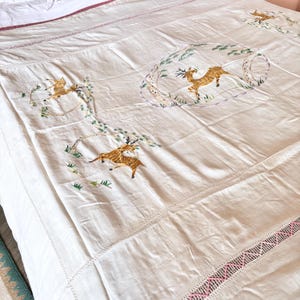 May include: Cream-colored embroidered bedspread featuring deer and foliage designs. The embroidery is in shades of brown, gold, green, and pink. The bedspread has a stitched border and is placed on a bed.