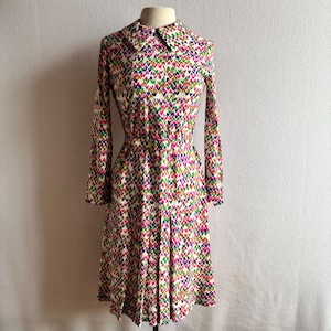 May include: A vintage dress with a colourful heart pattern. The dress has long sleeves, a collared neckline, and a pleated skirt. The pattern includes pink, green, purple, and blue hearts on a cream background. The dress is displayed on a mannequin.
