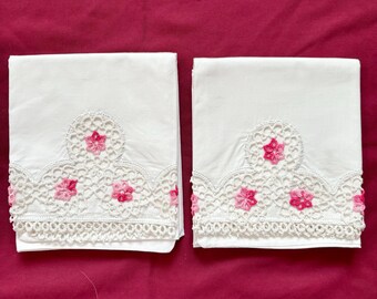 May include: Two white pillowcases with delicate lace trim and pink floral embroidery. The lace features a scalloped edge and small pink flowers.
