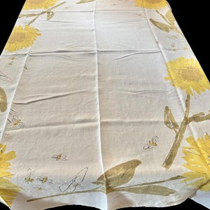 Könnte beinhalten: Weißes Tischtuch mit einem sich wiederholenden Muster aus gelben Sonnenblumen und grünen Stielen. Das Design beinhaltet kleine Bienen. Das Tischtuch ist aus hellem Stoff.