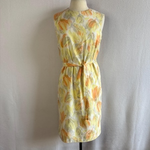 May include: A sleeveless, knee-length dress with a floral pattern in yellow, orange, and grey. The dress has a matching belt cinching the waist. The fabric appears lightweight, suitable for warmer weather.