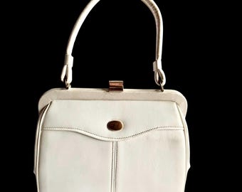 May include: A structured white handbag featuring a single top handle, a gold-toned metal clasp closure at the top, and a small oval gold-toned emblem centered on the front flap. The bag has a clean, minimalist design with visible stitching.