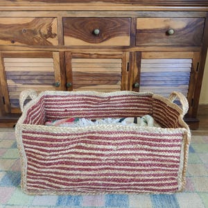 May include: A rectangular storage basket crafted from braided natural jute fibres, featuring horizontal stripes in alternating shades of tan and deep red. The basket includes two integrated side handles and a sturdy, woven construction.