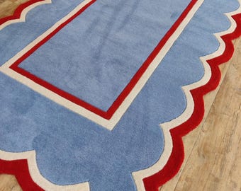May include: A rectangular area rug featuring a light blue center, a cream-colored border, and a vibrant red scalloped edge. The rug has a tufted texture and a layered design with a central rectangular panel framed by a thick cream and red border.
