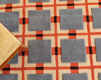 Op de afbeelding: Een tapijt met een ruitpatroon van oranje en zwarte lijnen op een beige achtergrond, met verhoogde grijze vierkante plukjes in elk rastergedeelte. Een houten hoekstuk met de tekst Warp & Weft is zichtbaar aan de linkerkant van het tapijt.