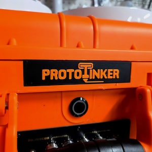Puede incluir: Maleta de color naranja brillante con la palabra "PROTO TINKER" en letras naranjas sobre una etiqueta negra. La maleta tiene un puerto circular negro y conectores plateados. La maleta está hecha de un material plástico duradero.