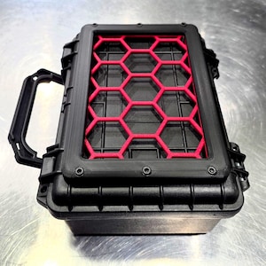 Può includere: Una custodia nera rettangolare con una griglia rossa a nido d'ape. La custodia ha una maniglia ed è fissata con viti. La custodia è progettata per proteggere gli oggetti dagli agenti atmosferici.