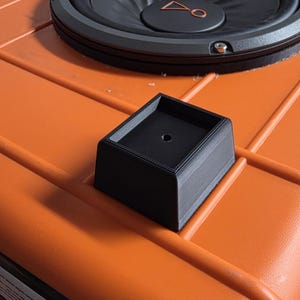 Peut inclure: Un objet noir imprimé en 3D de forme carrée avec un trou central, posé sur une surface orange. Un haut-parleur noir est visible en arrière-plan. L'objet semble être un petit support.