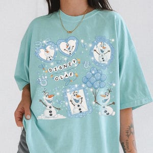 May include: A light blue t-shirt featuring a Disney Olaf design. The design includes multiple images of Olaf, the snowman character, in various poses, along with the text "Disney Olaf". The design is surrounded by hearts, butterflies, and balloons.