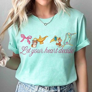 Peut inclure: T-shirt bleu clair avec un n&oelig;ud rose, une lampe dor&eacute;e, un singe avec une trompette et un tapis volant. Le texte "Let your heart decide" est &eacute;crit en rose. Col rond et manches courtes.