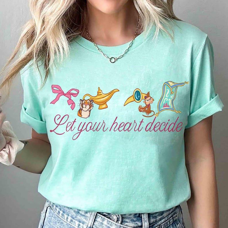 Peut inclure: T-shirt bleu clair avec un n&oelig;ud rose, une lampe dor&eacute;e, un singe avec une trompette et un tapis volant. Le texte "Let your heart decide" est &eacute;crit en rose. Col rond et manches courtes.