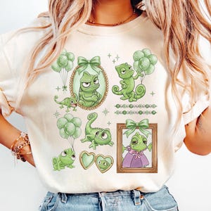 May include: Cream-colored t-shirt featuring a collection of green chameleon illustrations. The designs include chameleons in frames, holding balloons, and with heart-shaped lockets. The illustrations are accented with green bows and stars.