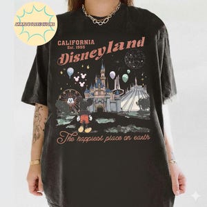 May include: Black t-shirt featuring a Disneyland graphic. The design includes the text "California Disneyland Est. 1955" and an illustration of the castle, Mickey Mouse, and other park elements. The phrase "The happiest place on earth" is also included.