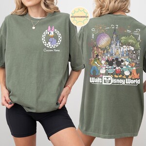 May include: Olive green t-shirt with a Daisy Duck graphic on the front, set within a checkered circle. The back showcases a colourful illustration of Walt Disney World characters and landmarks, with the words "Walt Disney World".