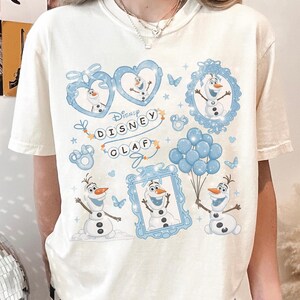 May include: Cream-colored t-shirt featuring a Disney Olaf graphic. The design includes multiple images of Olaf the snowman in blue frames, hearts, and holding balloons. The text "Disney Olaf" is also printed on the shirt.