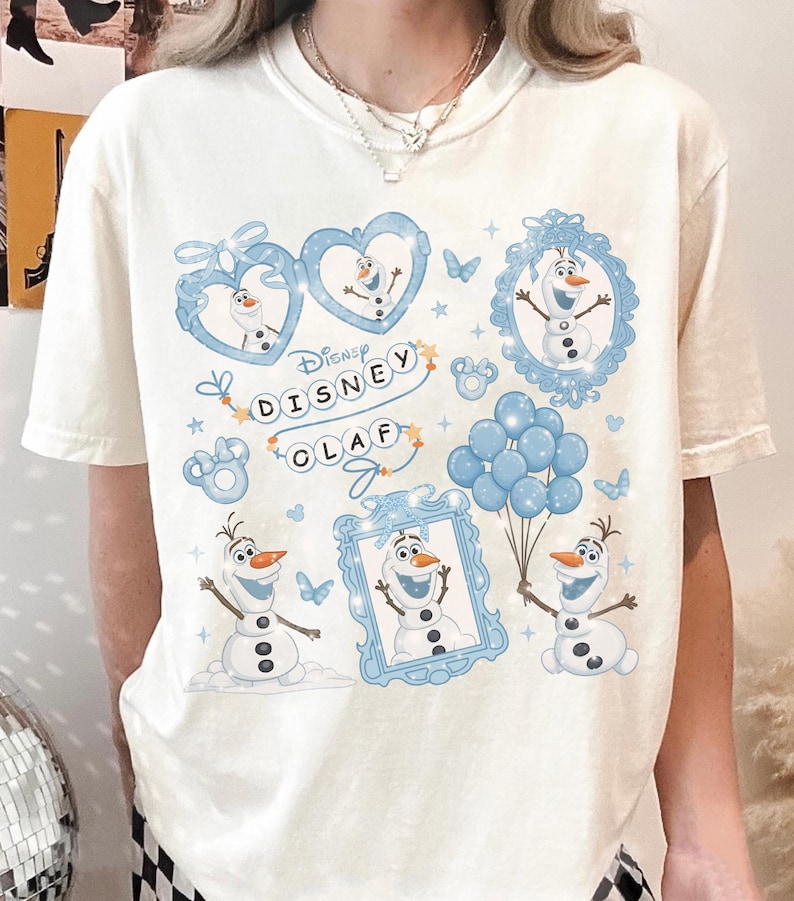 May include: Cream-colored t-shirt featuring a Disney Olaf graphic. The design includes multiple images of Olaf the snowman in blue frames, hearts, and holding balloons. The text "Disney Olaf" is also printed on the shirt.
