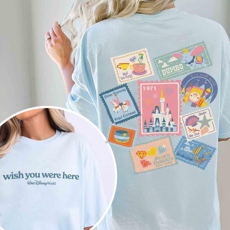 May include: Light blue t-shirt with the text "wish you were here" and various Disney-themed stamp designs on the back. The stamps feature Dumbo, a carousel, a castle, and other park attractions.