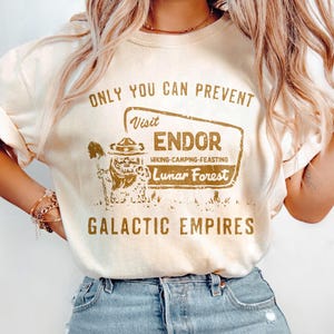 May include: Cream-coloured sweatshirt with a brown graphic and text that reads "ONLY YOU CAN PREVENT Visit ENDOR Lunar Forest GALACTIC EMPIRES." The graphic features a bear-like character in a ranger hat.