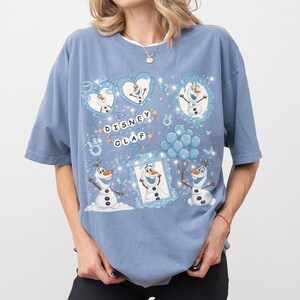 May include: A light blue t-shirt featuring a Disney Olaf graphic. The design includes multiple images of Olaf, the snowman character, within frames and hearts, along with the text "Disney Olaf". The shirt has short sleeves and a relaxed fit.