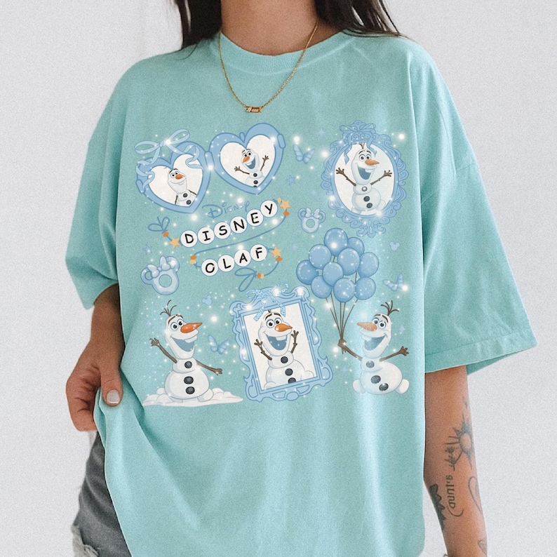 May include: A light blue t-shirt featuring a Disney Olaf design. The design includes multiple images of Olaf, the snowman character, in various poses, along with the text "Disney Olaf". The design is surrounded by hearts, butterflies, and balloons.