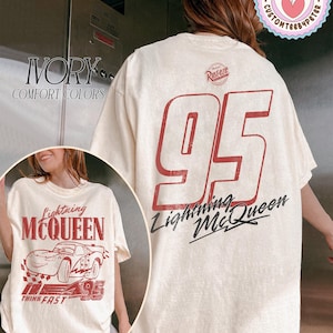 May include: Ivory-colored Lightning McQueen t-shirt. The front features a graphic of the character and the text "Lightning McQueen" and "95". The back displays a large "95" and "Lightning McQueen" in red and black.