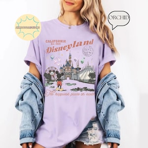 May include: A light purple t-shirt featuring a Disneyland graphic. The design includes the Disneyland logo, a castle, Mickey Mouse, and the phrase "The happiest place on earth." The shirt is a relaxed fit.