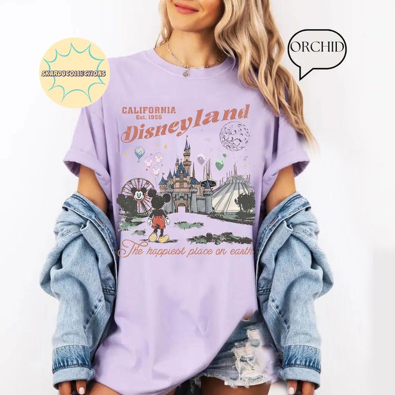 May include: A light purple t-shirt featuring a Disneyland graphic. The design includes the Disneyland logo, a castle, Mickey Mouse, and the phrase "The happiest place on earth." The shirt is a relaxed fit.