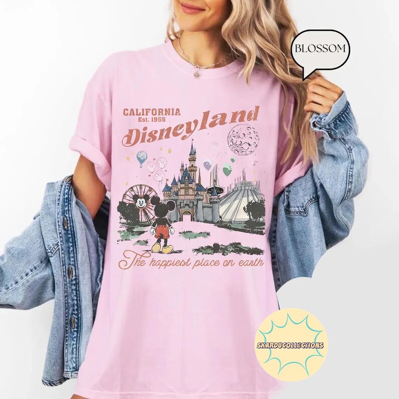 May include: Pink t-shirt with a Disneyland graphic. The design features a Ferris wheel, castle, and Mickey Mouse, with text reading "California Disneyland Est. 1955" and "The happiest place on earth."