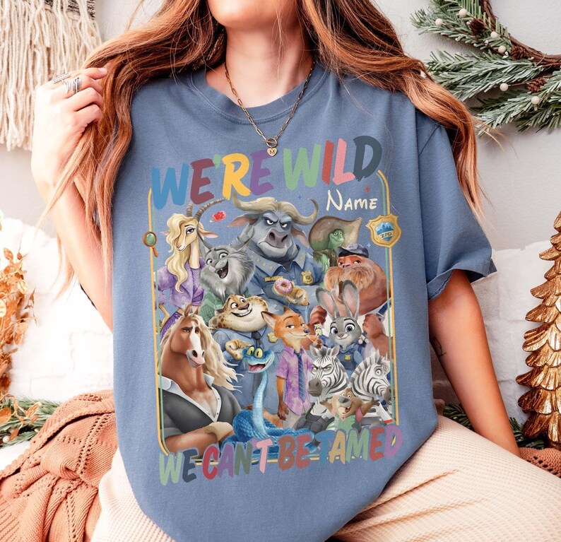 May include: A blue t-shirt featuring a colorful graphic of various cartoon animal characters. The text "WE'RE WILD" is at the top, with space for a name, and "WE CAN'T BE AMED" at the bottom. The design includes a badge.