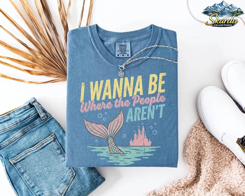 Puede incluir: Camiseta azul con el texto "I Wanna Be Where the People Aren't" y un gr&aacute;fico de cola de sirena. Un collar plateado y zapatos blancos est&aacute;n al lado de la camisa. Unos vaqueros azules y una bufanda beige tambi&eacute;n est&aacute;n en la imagen.
