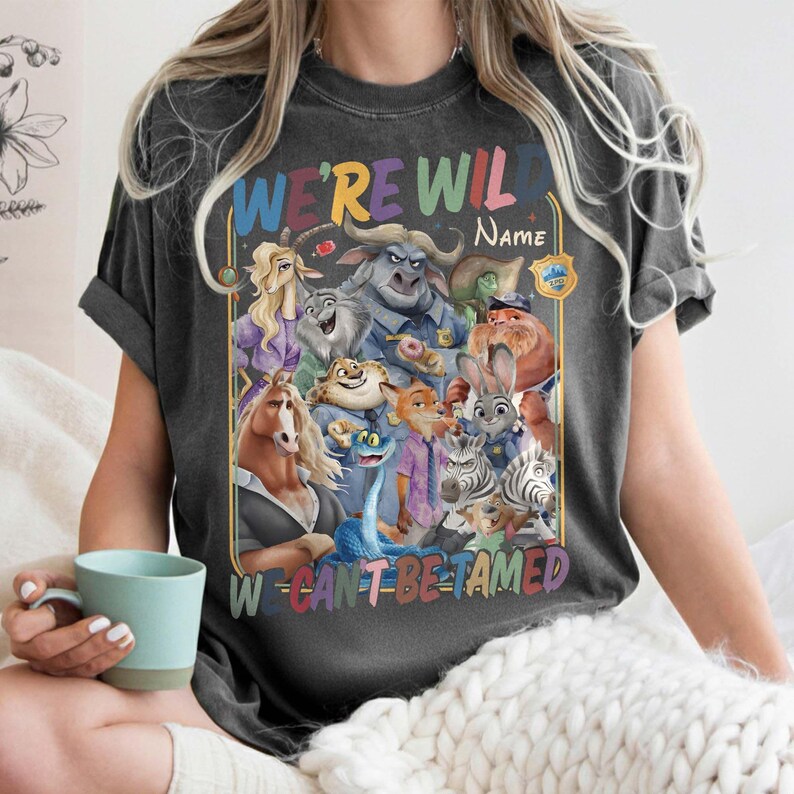 May include: Dark gray t-shirt featuring a colorful graphic of animated animal characters. The text "WE'RE WILD" and "WE CAN'T BE TAMED" is displayed in rainbow colors. A name can be added to the design.