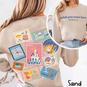 May include: Sand-colored sweatshirt featuring a collection of colorful Disney-themed postage stamp designs on the back. The stamps depict various attractions and characters, including Dumbo, a carousel, and a castle. The front of the sweatshirt says "wish you were here".