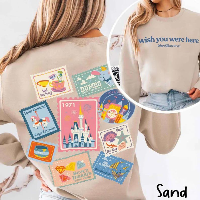 May include: Sand-colored sweatshirt featuring a collection of colorful Disney-themed postage stamp designs on the back. The stamps depict various attractions and characters, including Dumbo, a carousel, and a castle. The front of the sweatshirt says "wish you were here".
