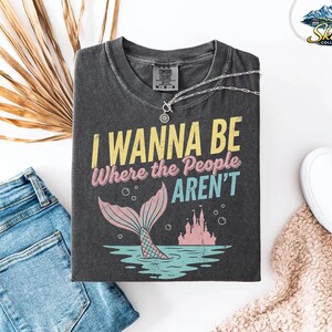 Puede incluir: Camiseta gris oscuro con el texto "I Wanna Be Where the People Aren't" en amarillo, rosa y turquesa. El dise&ntilde;o incluye una cola de sirena, burbujas y un castillo. Un collar plateado est&aacute; sobre la camiseta, con vaqueros azules y zapatos blancos cerca.