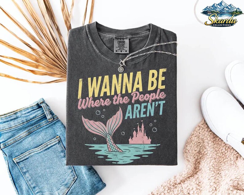 Puede incluir: Camiseta gris oscuro con el texto "I Wanna Be Where the People Aren't" en amarillo, rosa y turquesa. El dise&ntilde;o incluye una cola de sirena, burbujas y un castillo. Un collar plateado est&aacute; sobre la camiseta, con vaqueros azules y zapatos blancos cerca.