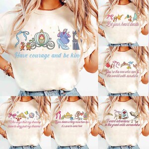 Peut inclure: Plusieurs t-shirts cr&egrave;me avec des motifs de contes de f&eacute;es. L'un d'eux pr&eacute;sente un carrosse et Cendrillon, avec le texte "Have courage and be kind". D'autres t-shirts affichent des phrases comme "Let your heart decide".