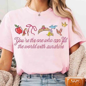Peut inclure: T-shirt rose clair avec un motif color&eacute; comprenant une pomme, un n&oelig;ud, une couronne et des oiseaux. Le texte "You're the one who can fill the world with sunshine" est imprim&eacute; en &eacute;criture cursive.