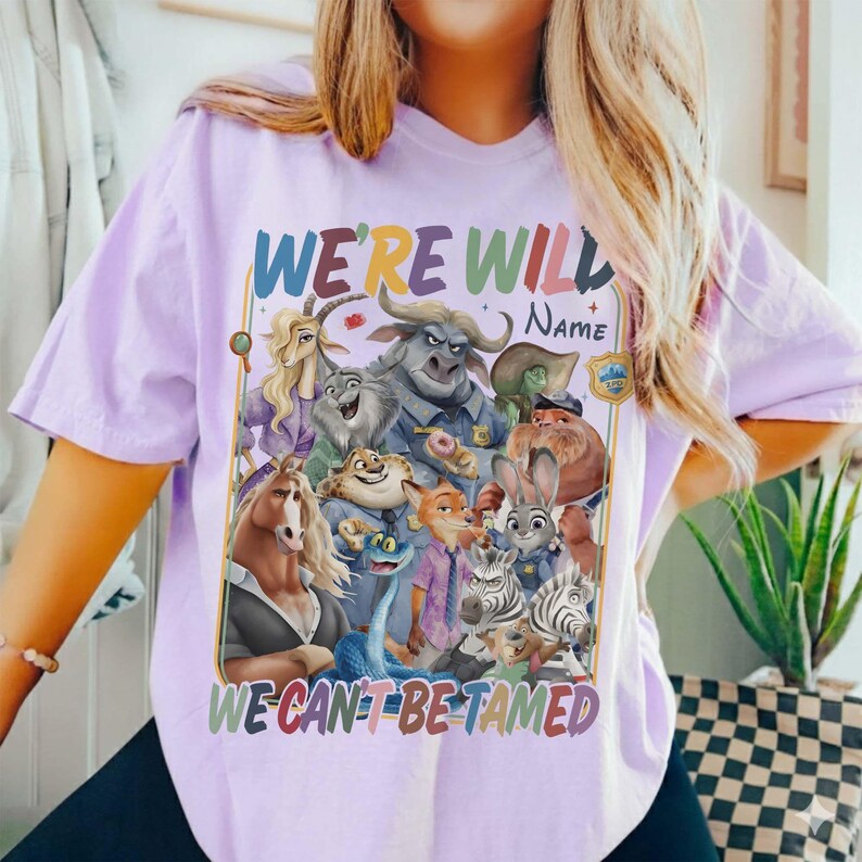 May include: A light purple t-shirt featuring a colorful graphic of various cartoon animal characters. The text "WE'RE WILD, WE CAN'T BE TAMED" is displayed above and below the characters in a rainbow font.