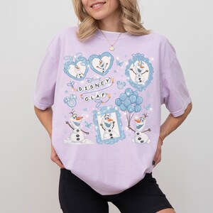 May include: A light purple t-shirt featuring multiple illustrations of Olaf from Disney's Frozen. The design includes Olaf in various poses, hearts, balloons, and the text "Disney Olaf". The shirt has short sleeves and a relaxed fit.
