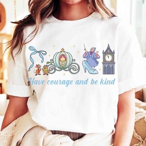 Peut inclure: T-shirt blanc avec une illustration sur le th&egrave;me de Cendrillon. Le motif comprend un carrosse, une tour d'horloge, une f&eacute;e marraine, des souris et le texte "Have courage and be kind" en bleu clair.