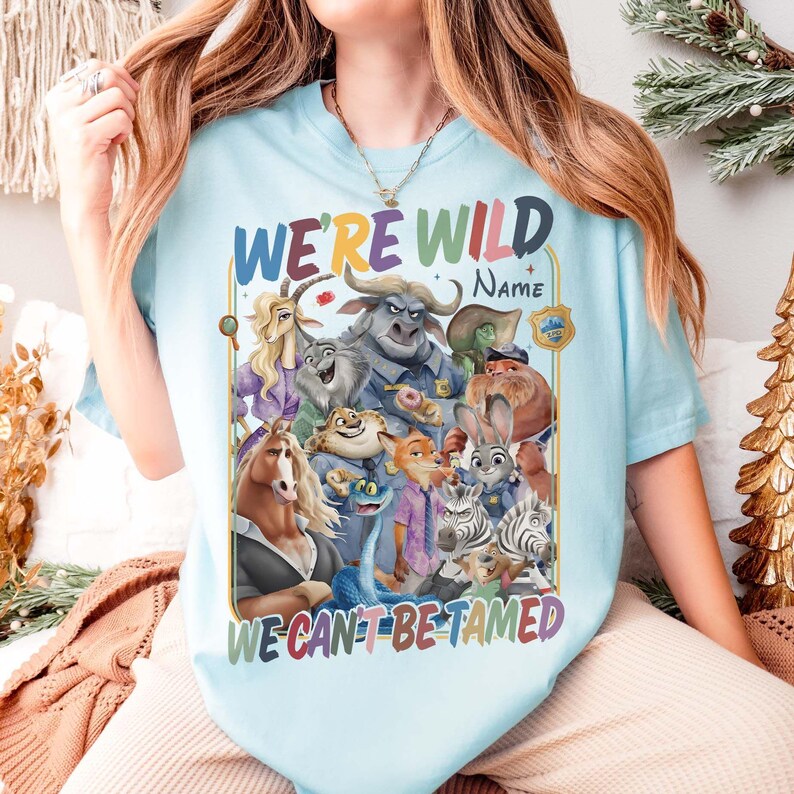 May include: Light blue t-shirt featuring a colorful graphic of various animal characters from a movie, with the text "WE'RE WILD" above and "WE CAN'T BE TAMED" below. The shirt has a crew neck and short sleeves.