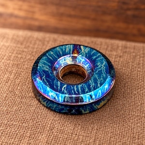 Puede incluir: Una cuenta metálica circular con un acabado iridiscente vibrante y un patrón azul y morado similar a un rayo. La cuenta tiene una superficie lisa y pulida con un orificio central, mostrando un brillo metálico que cambia de color.