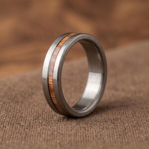 Puede incluir: Un anillo de metal plateado cepillado con una incrustación central de madera de color marrón claro. La banda tiene un perfil redondeado y liso con un acabado metálico que contrasta con la textura natural de la madera en el centro.
