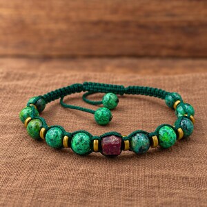May include: A macrame bracelet featuring round green speckled beads and small metallic gold-toned spacers. A single rectangular deep red stone bead is positioned at the center. The bracelet is secured with a dark green adjustable cord closure.