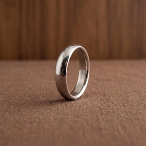 May include: A polished silver-toned metal ring with a smooth, rounded exterior surface and a plain, unadorned band design. The ring features a reflective finish that catches the light, showcasing a simple, circular shape standing upright on its edge.