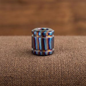 Puede incluir: Una cuenta cilíndrica con acabado metálico y rayas verticales en tonos azul, cobre y plata. La cuenta tiene una superficie lisa y pulida con un orificio central que atraviesa la parte superior e inferior, mostrando un degradado multicolor.